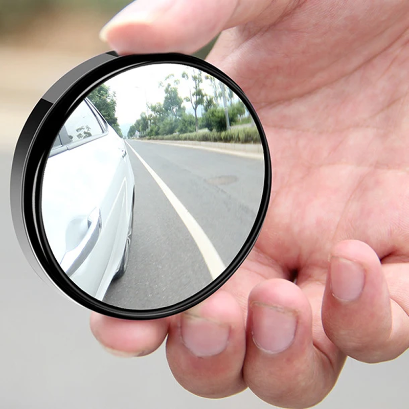 Hand holding a small round mirror reflecting a road scene with trees.