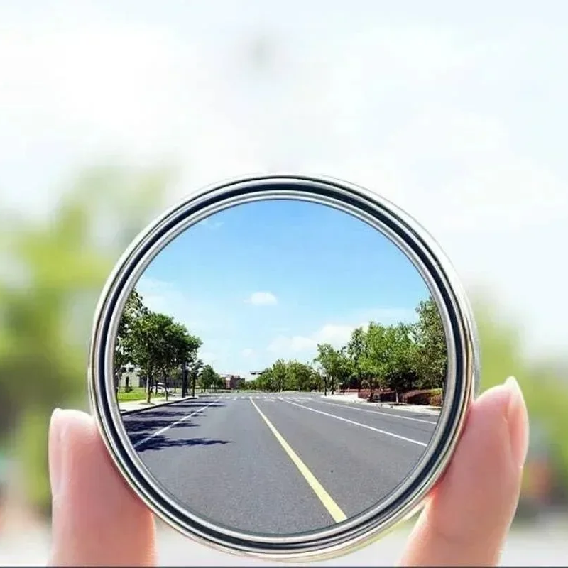 Round mirror held by a hand reflecting a road with trees and blue sky.
