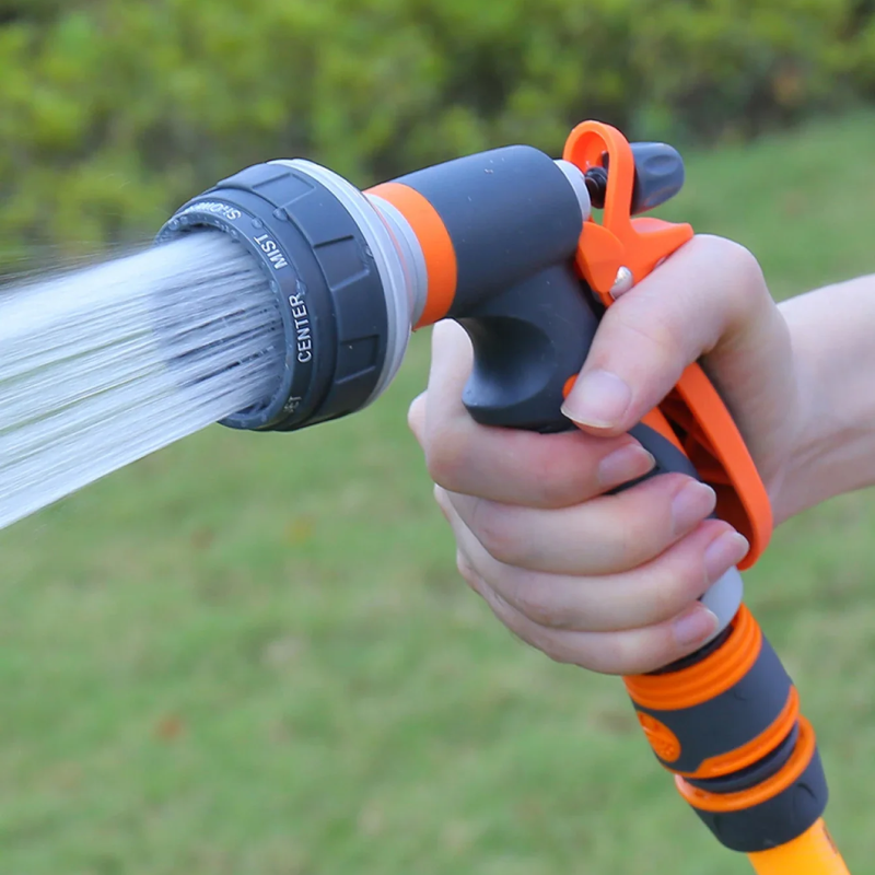 Hand holding an orange and gray garden hose nozzle with water spraying outdoors.