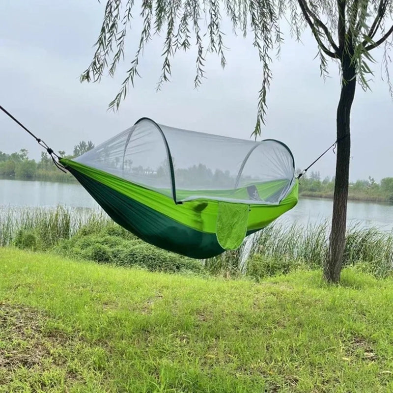 Green hammock with mosquito net hanging between two trees by a body of water.